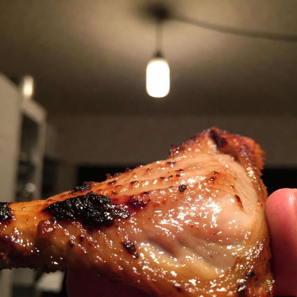 Charred chicken skin under bulb, shaky thumb intrudes.