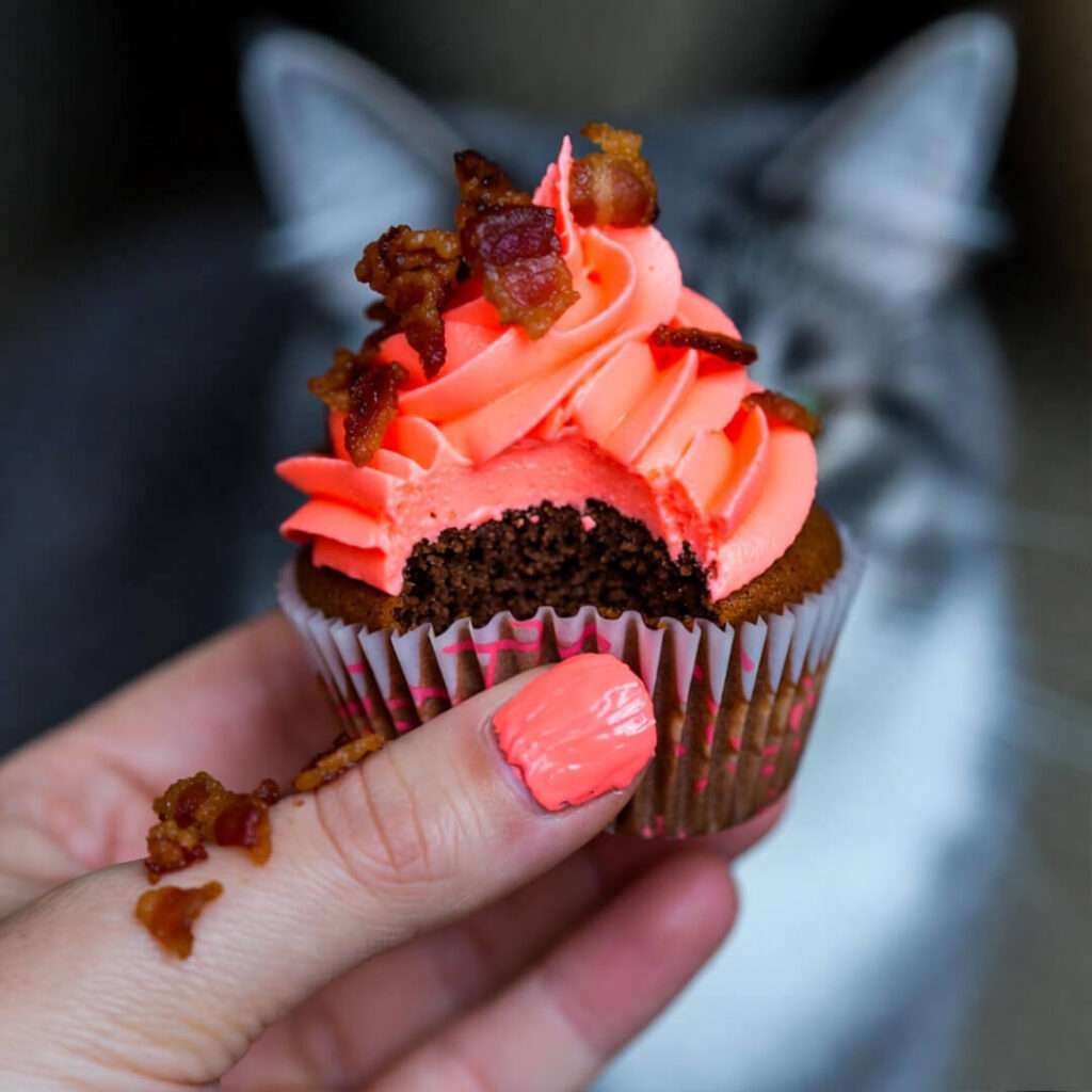 Frosting-smeared thumb holds bacon cupcake, cat glares.